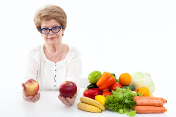 Attractive elder Caucasian woman holding two red apples, fresh vegetables and fruits are on table, white background