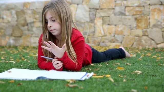 Blond kid girl maths homework laying on grass counting with fingers writing in notebook