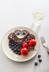 On the table there is a plate filled with chocolate cakes, blueberries, tomato snacks, fruit, a glass of lemonade