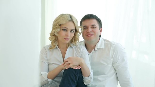 Husband and wife sitting on the floor next to each other near a large window, they hug, looking at the camera.Happy family in white shirts and jeans. Man and woman smiling, hugging.