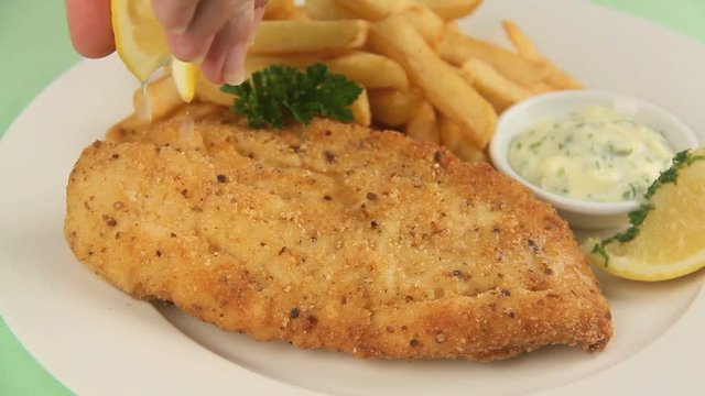 Squeezing fresh lemon over a fried fish fillet with hot chips and tartare sauce garnished with parsley.
