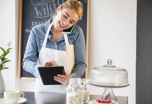 Caucasian barista using cell phone and digital tablet in cafe - Powered by Adobe