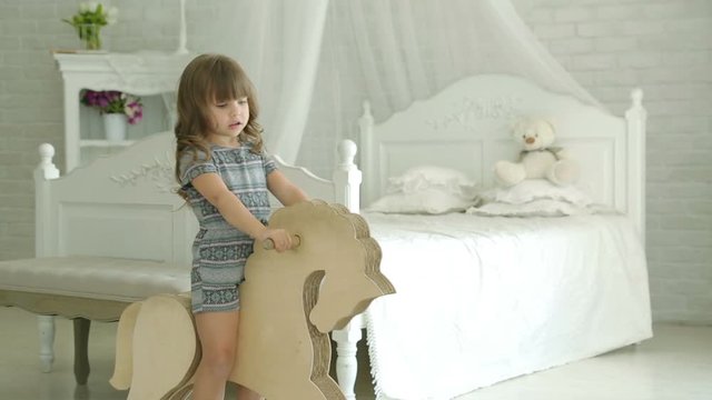 A little girl riding on wooden horse. Girl smiles. The little girl in the bedroom against a white four-poster beds. On the background of white brick wall and a chest of drawers. Bedroom.