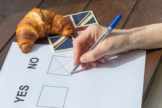 Female hand filled NO in the printed form. On the wooden table is lying a napkin English flag design, on which is symbolically lying a French croissant.