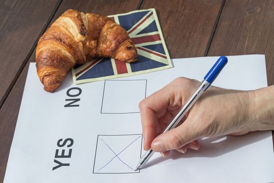Female hand filled YES in the printed form. On the wooden table is lying a napkin English flag design, on which is symbolically lying a French croissant.