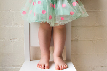 Toddler girl in green and pink polka dot skirt standing on white chair against white brick wall (cropped)