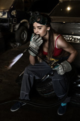 Girl mechanic sitting on a tire with blowpipe and smoking in a garage. car repairs. colorless life concept