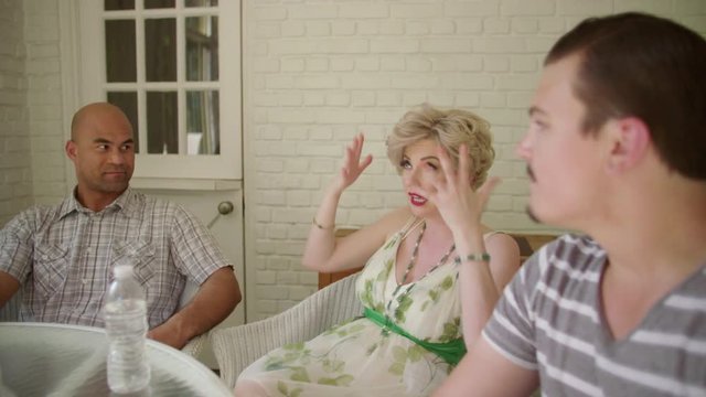 Attractive blonde woman explains something using gestures to a group of friends on a sunny indoor porch.  Focus changes to different people throughout shot.  Hand held camera