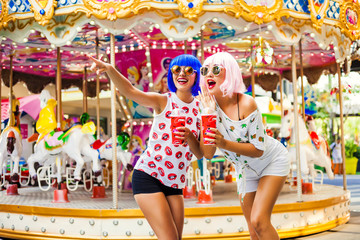 Two beautiful girls in bright colored wigs to cocktails in hand having fun at amusement park