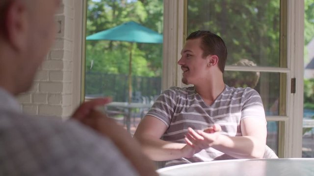 Attractive blonde woman and dark-haired man sit, talking and laughing in covered porch, on a summer day.  Hand held mid shot, originally recorded in slow motion at 60fps.  Tinted