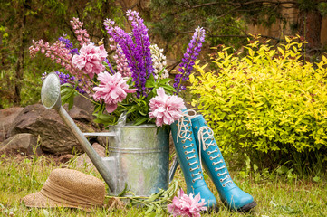Gardening tools and a straw hat on the grass in the garden. Watering can and blue rubber boots