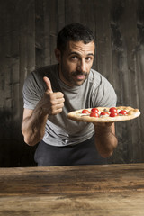 Ragazzo con i capelli scuri con la pizza in mano su sfondo legno scuro  su un tavolo di legno fa il segno ok.