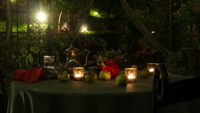 Flickering candles, wild apples and tea glasses on a table that is under a gazebo. Cozy, intimate, romantic lighting in the evening with the camera sliding toward the table. Dolly shot.