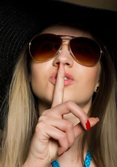 Close-up of attractive brunette woman in a bikini, big hat and sunglasses putting a finger on her...