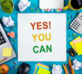 Yes You Can. Office table desk with supplies, white blank note pad, cup, pen, pc, crumpled paper, flower on blue background. Top view