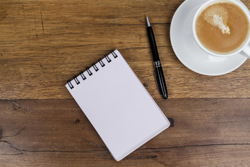 White sheets notebook with spiral in the center of the wooden dark brown table with black pen aside and cup of coffee on top right from above