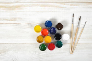 Three paint brushes and colorful gouache containers on wooden surface in top view.