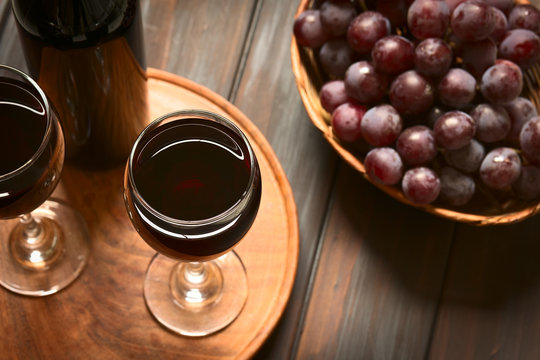 Glasses of red wine with red globe grapes and a bottle of wine, photographed on dark wood with natural light (Selective Focus, Focus on the rim of the wine glass)