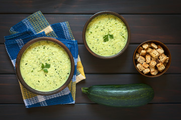 Cream of zucchini soup garnished with parsley leaf, with a bowl of homemade croutons and a raw zucchini on the side, photographed on dark wood with natural light