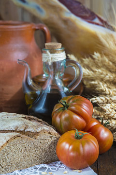 Bodegón de comida con aceite de oliva, pan, tomates, jamón y espigas de cereales en ambiente rústico