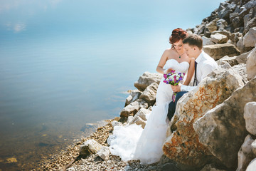 Happy smiling young bride and groom, walking on the rocks, beautiful sky, water.