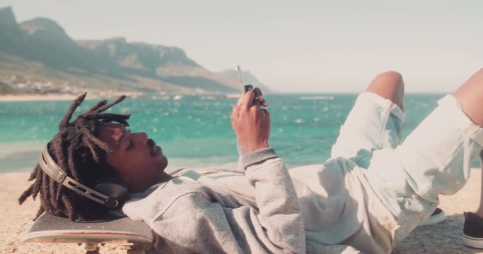 African American Skater Resting and Looking at Smartphone at Sea