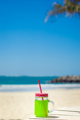 Fototapeta na wymiar Vertical photo of green juice in a funny glass with a red straw, beach as background