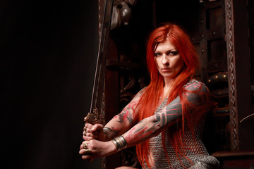Studio shot of beautiful young redhead girl with multiple tattoo in chainded armour with the sword