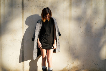Stylish young girl dressed in a long black T-shirt with a coat thrown over shoulders posing against a background of a concrete wall