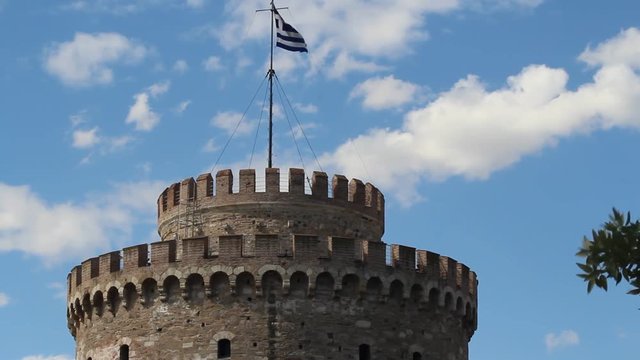 White Tower in Thessaloniki, Greece. Greek flag waving on the top of the tower in front of some clouds in a blue sky.