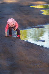 Little girl bending to the ground playing in big spring puddle wearing purple nylon and pink bucket hat 