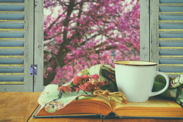 romantic scene of cup of coffee next to old book in front of countryside view outside of the old rustic window. vintage filtered and toned image
