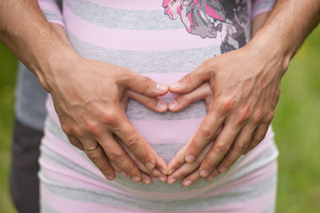 Closeup shot of pregnant woman with heart from her and her man hands on her belly dressed in striped pink and grey dress