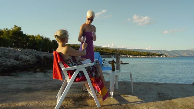 Two female enjoy and relaxing on pier in the sunset time, with a glass of wine in hand, watching the sea. View from side and panning in to the sea.
