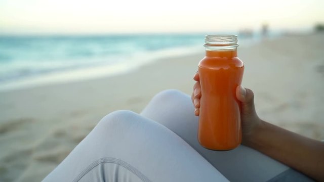 Cold pressed juice - close up of woman drinking carrot juice on beach living healthy active life. Fit girl enjoying healthy diet and nutrition after yoga at beach. 