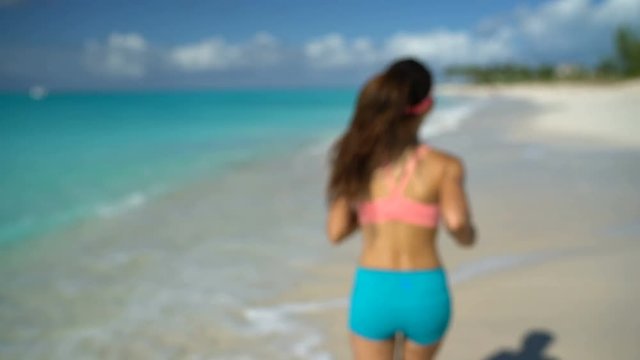 Runner listening to music running exercising on beach. Young woman putting headphones in ear by sea shore. Female fitness girl training listening music while jogging on sunny day. 