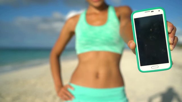 Empty Blank phone screen. Young sporty woman showing smart phone with empty screen on beach. Female fitness girl is wearing green sports bra. Fit lady is standing in nature.