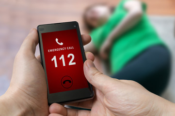 Man dialing emergency (112 number) on smartphone. Woman had heart attack and is lying on the floor. - obrazy, fototapety, plakaty