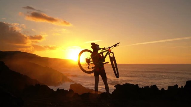 Success, achievement, accomplishment and winning concept with cyclist mountain biking. Happy MTB woman cycling raising arms lifting bike by sea during sunset cheering and celebrating at summit top. 