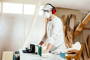 Carpenter working on wood machine in a woodworking factory, wearing face mask and headphones for noise