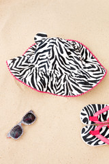 A set of beach accessories in black and white stripes with pink details. Sunglasses, Flip Flop and Sunhat on the sand closeup