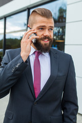 Young bearded businessman using mobile phone outside the office.