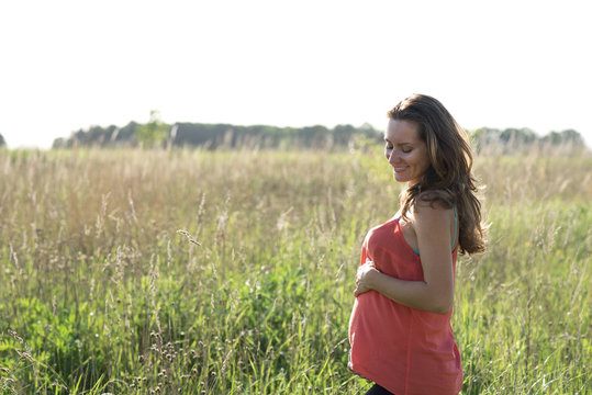Young beautiful pregnant woman holding tummy smiling, in red a light summer dress, happy on meadow the grass 