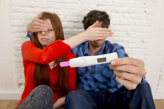 young sad couple  in shock and surprise holding pink positive pregnancy test blocking eyes scared