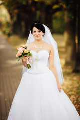 Fototapeta na wymiar Portrait of beautiful young brunette bride in elegant dress with bouquet of pink flowers in the autumn park