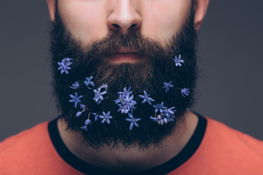 Close-up of young shirtless man with flowers in his beard standing against grey background