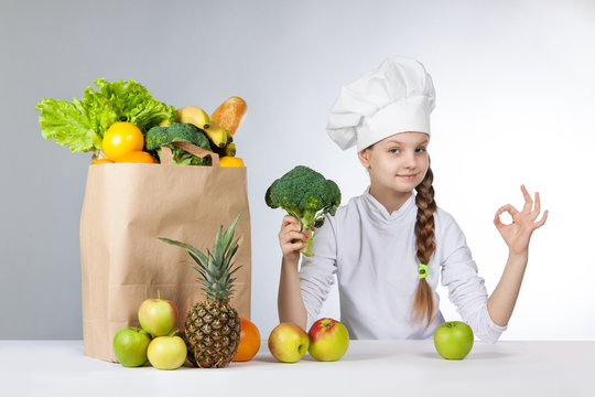 Little happy girl in a cap cook a variety of fresh food. Girl holding broccoli. and shows OK. Positive human emotion, facial expression feeling, attitude. Bag with fresh vegetables and fruits.