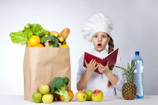Little girl in a cap cook a variety of fresh food. Girl holds red book in hands. Wonder human emotion, facial expression feeling