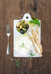 Fresh feta cheese with olives, basil, rosemary and bread slices on white ceramic serving board over rustic wooden background