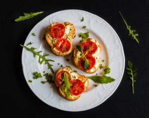 Toasts (Crostini) with ricotta, cherry tomatoes and arugula on black background. Top view.